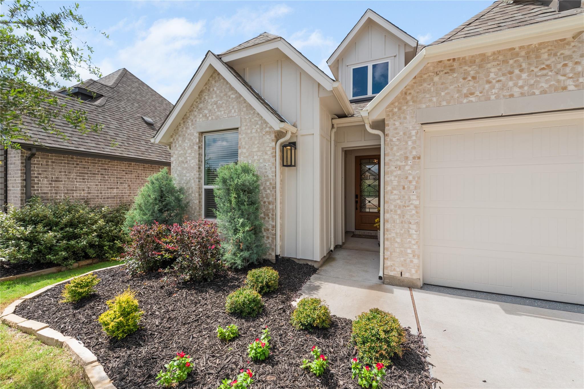 Light brick home entrance with a covered walkway, white garage door, and landscaped flower beds.