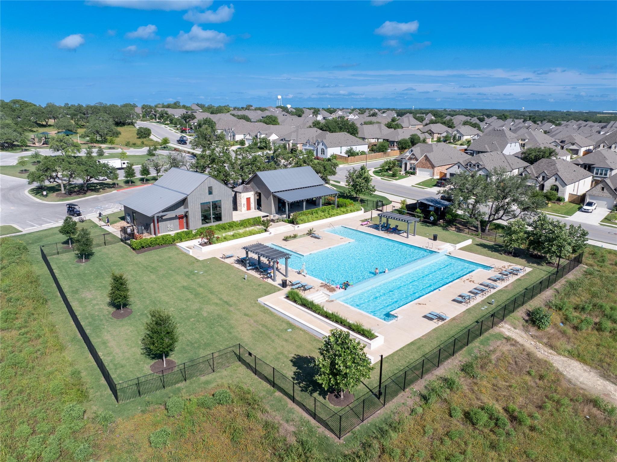 Aerial view of the neighborhood pool, clubhouse, shaded seating, and fenced grassy grounds.