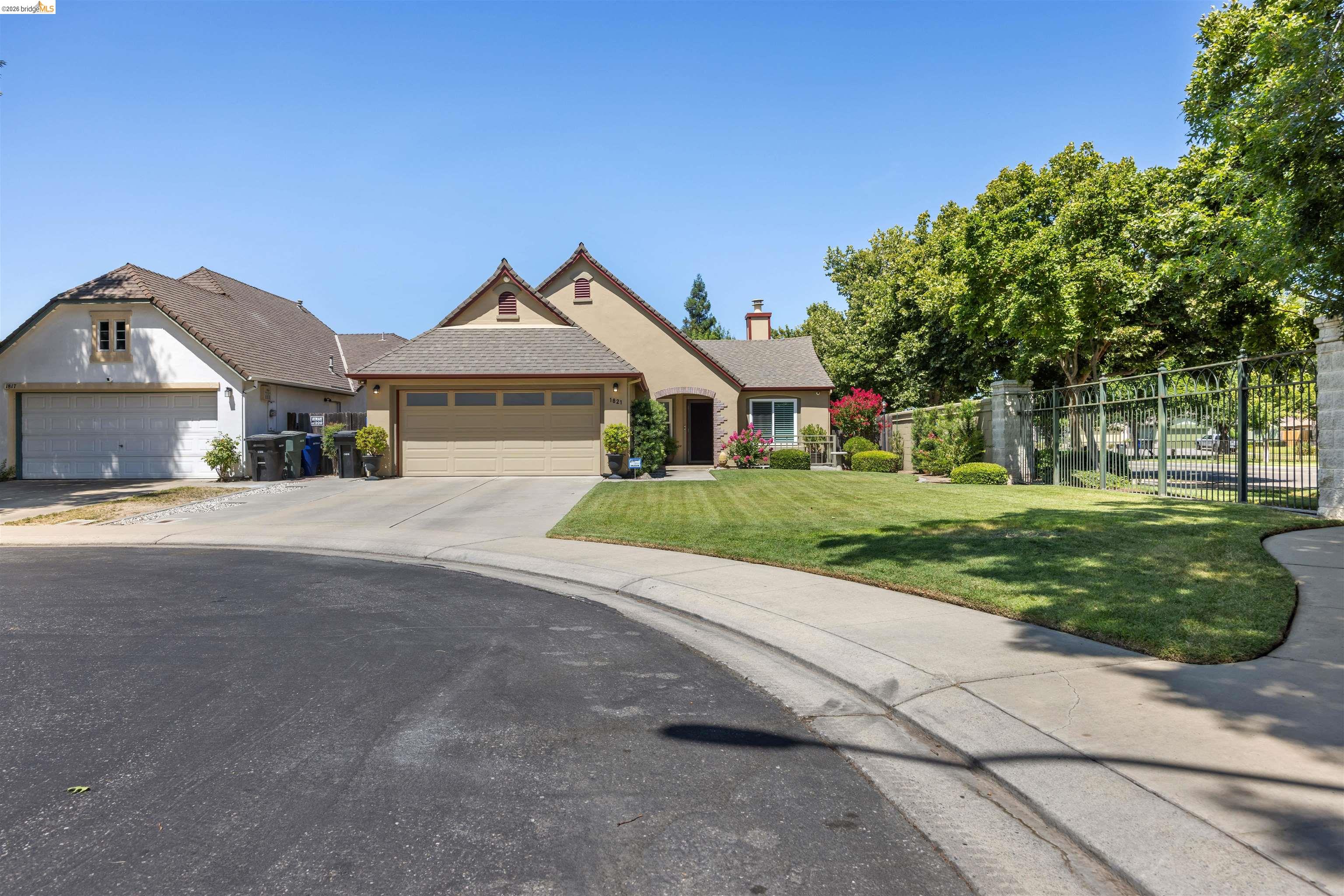 Tan stucco home with a two-car garage, landscaped front lawn, and mature shade trees.