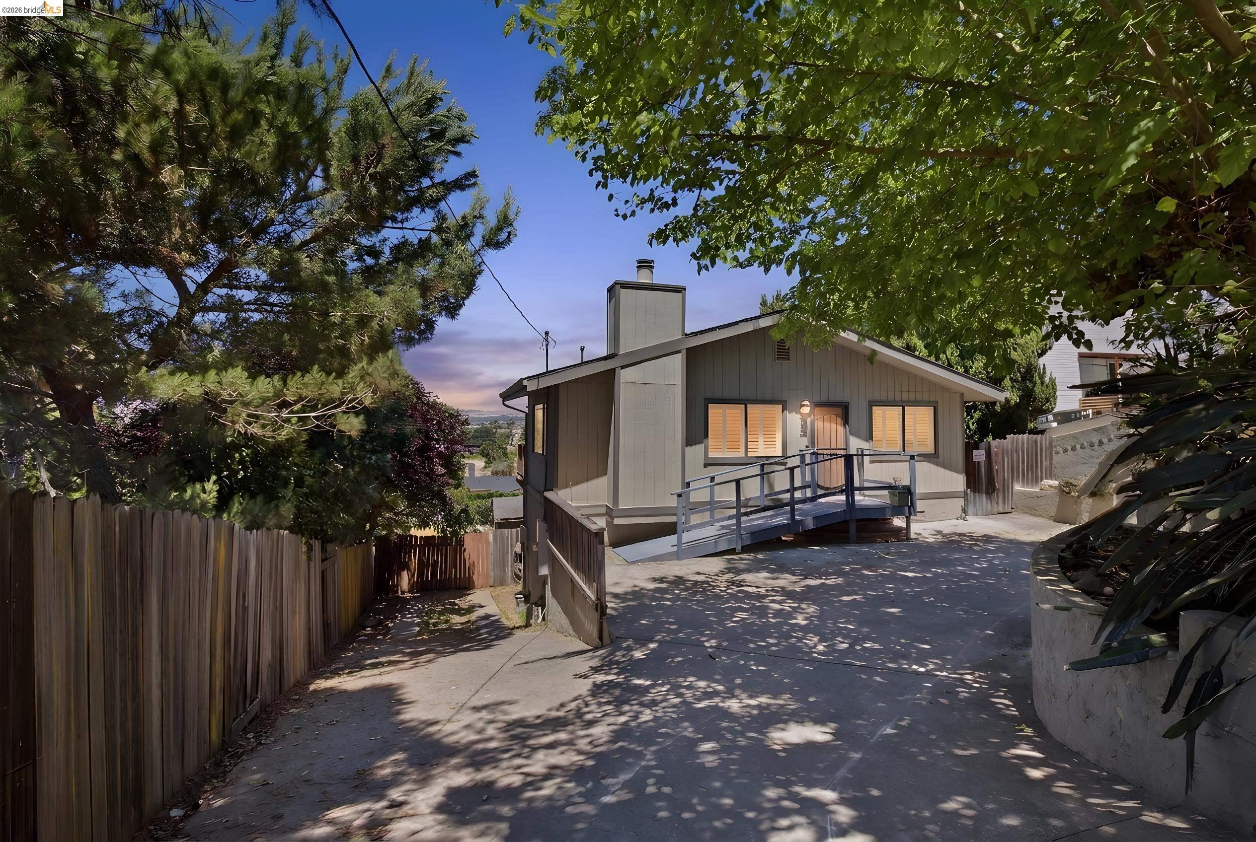Gray single-story home with a wheelchair-accessible ramp, shaded driveway, and wooden fence.