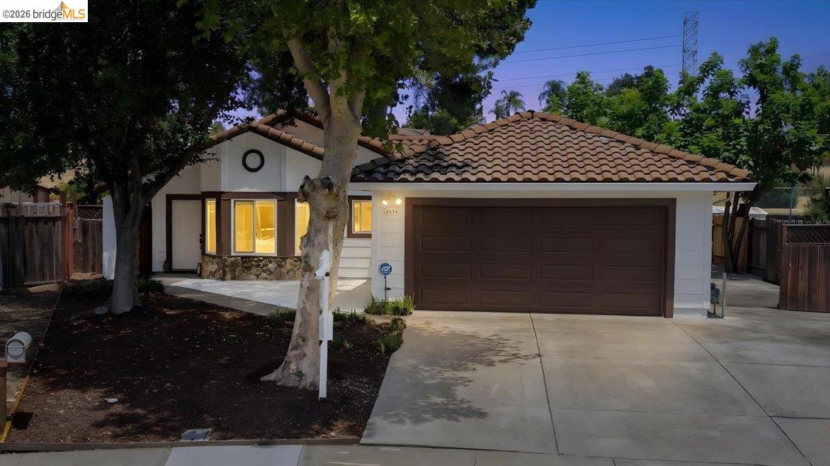 Single-story white house with a brown tile roof, dark double garage door, and mature trees in front.