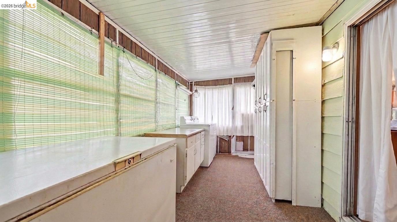 Narrow laundry room with side counters and cabinets, window blinds, and a washer at the far end.