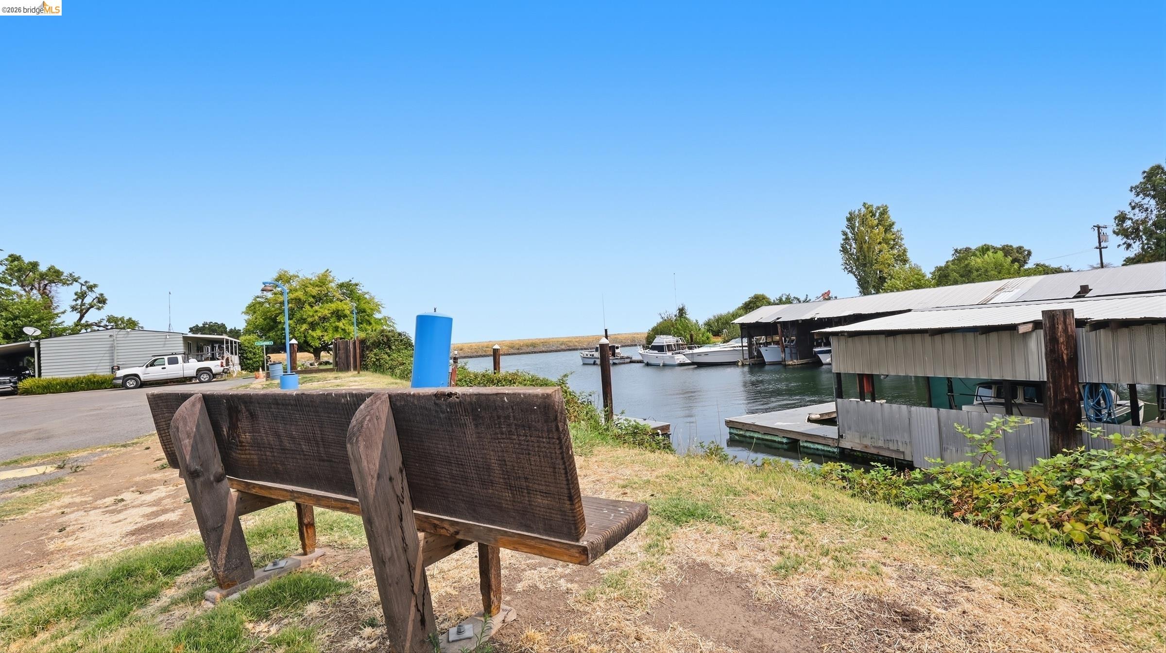 Waterfront marina with covered boat slips, boats, and a large wooden bench in the foreground.