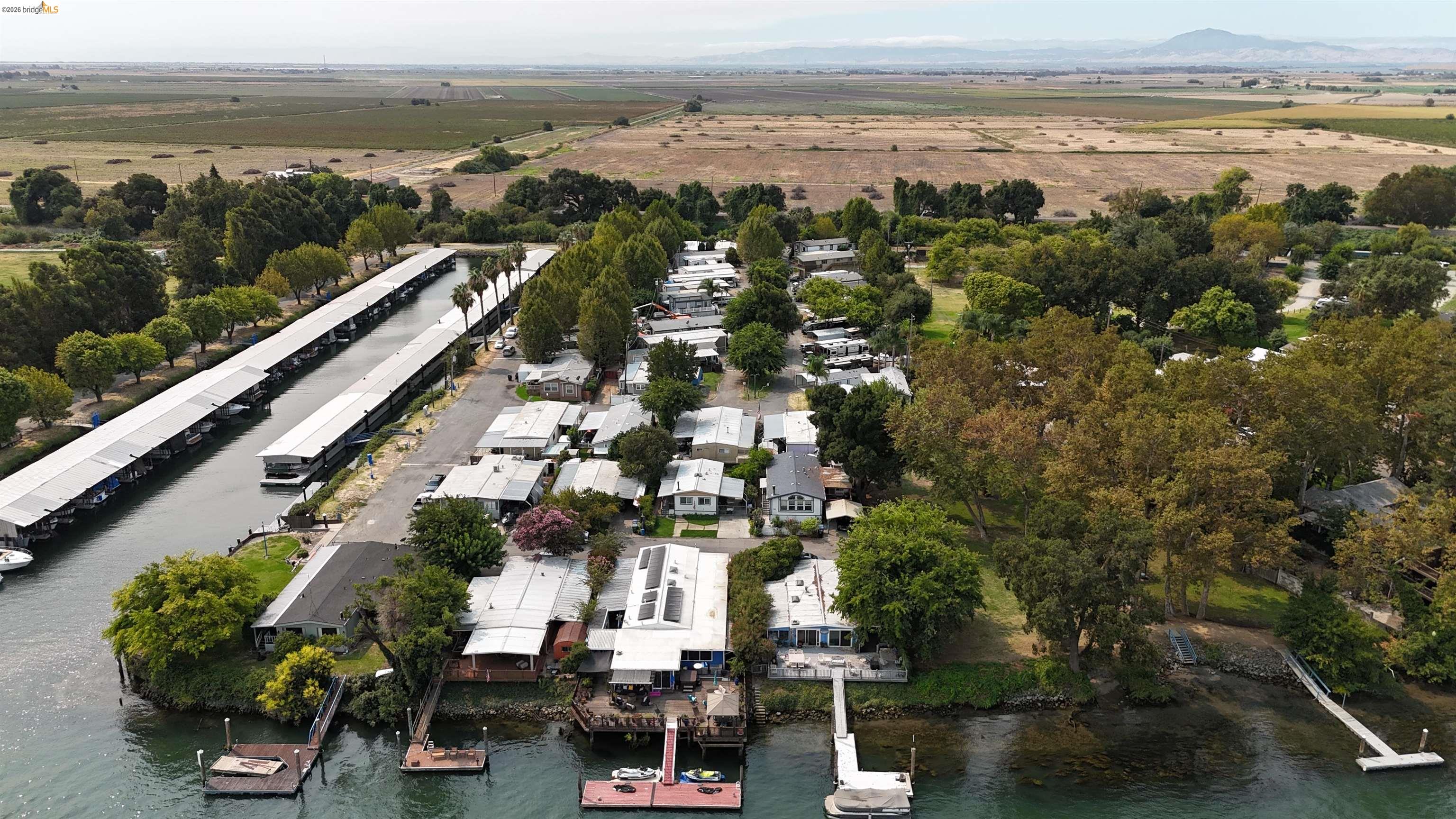 Waterfront homes and covered boat slips line a canal, viewed from above with fields beyond.
