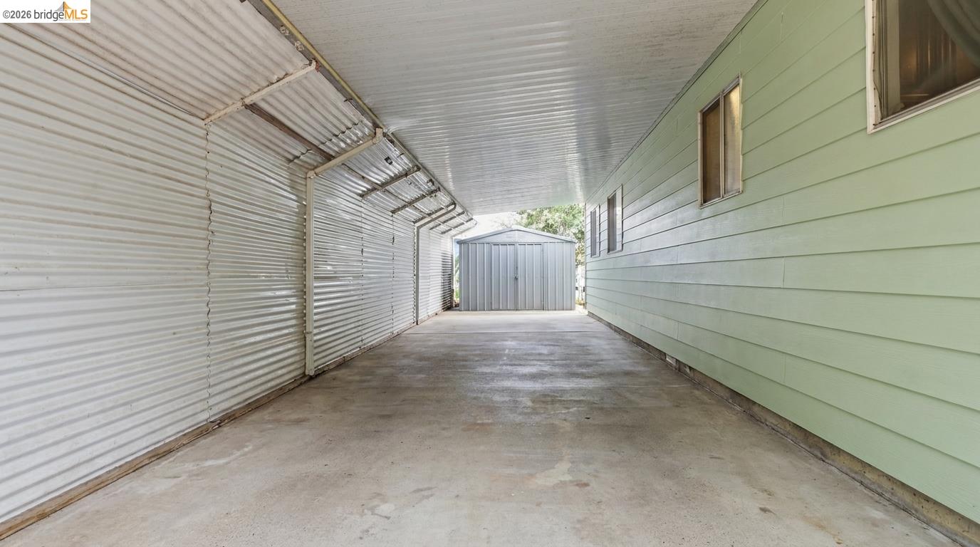 Covered concrete driveway beside a light-green house, ending at a metal storage shed.