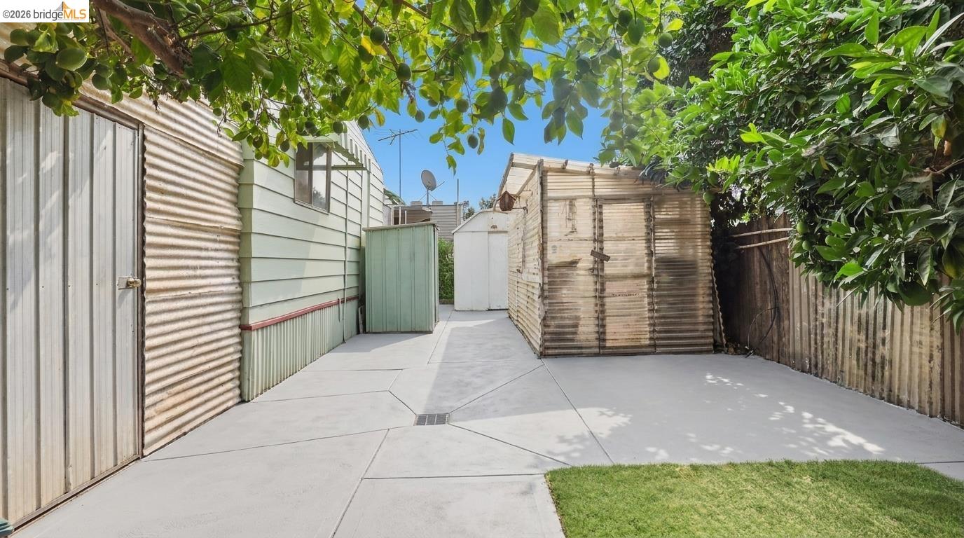 Paved backyard courtyard with leafy shade trees, sheds, and a small patch of lawn.