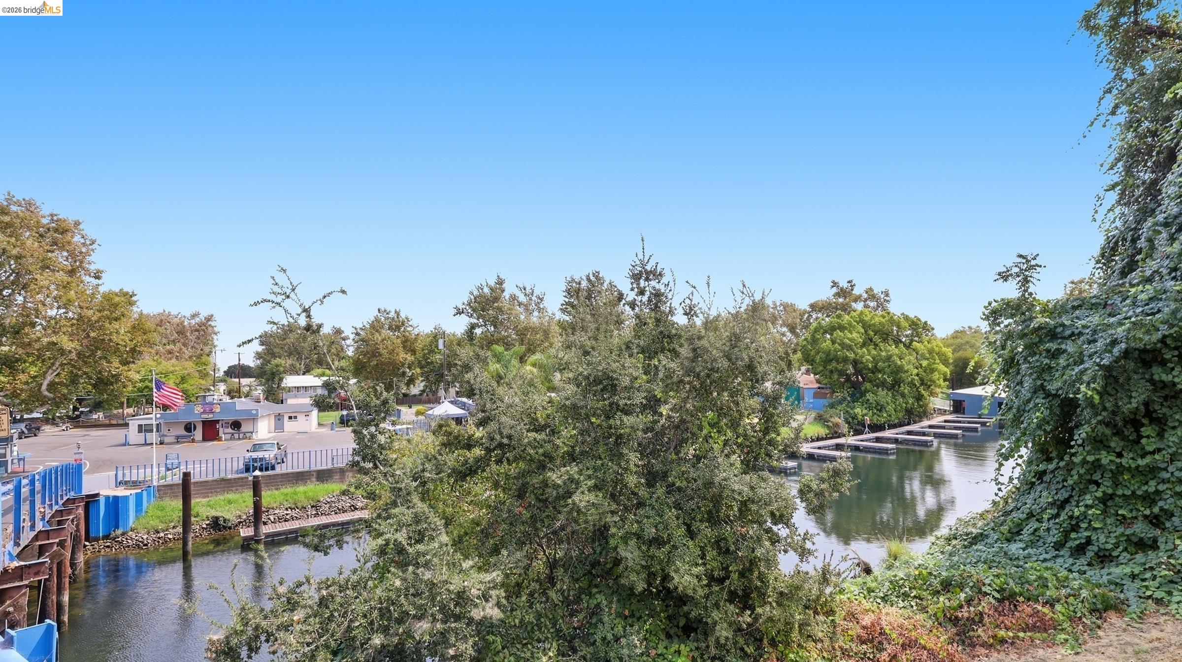 Tree-lined canal with docks and waterfront buildings beneath a clear blue sky.