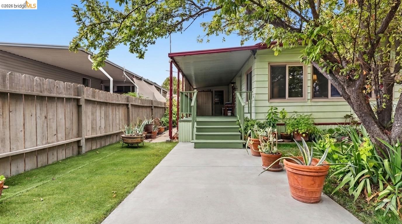Green house’s covered rear porch and steps beside a concrete walkway, potted plants, and a fenced lawn.