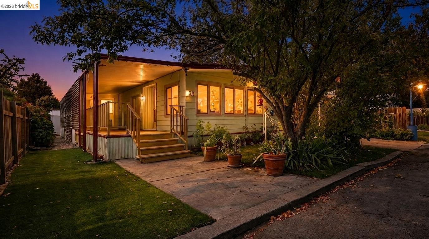 Single-story home at dusk with a warmly lit covered porch, paved patio, and large shade tree.