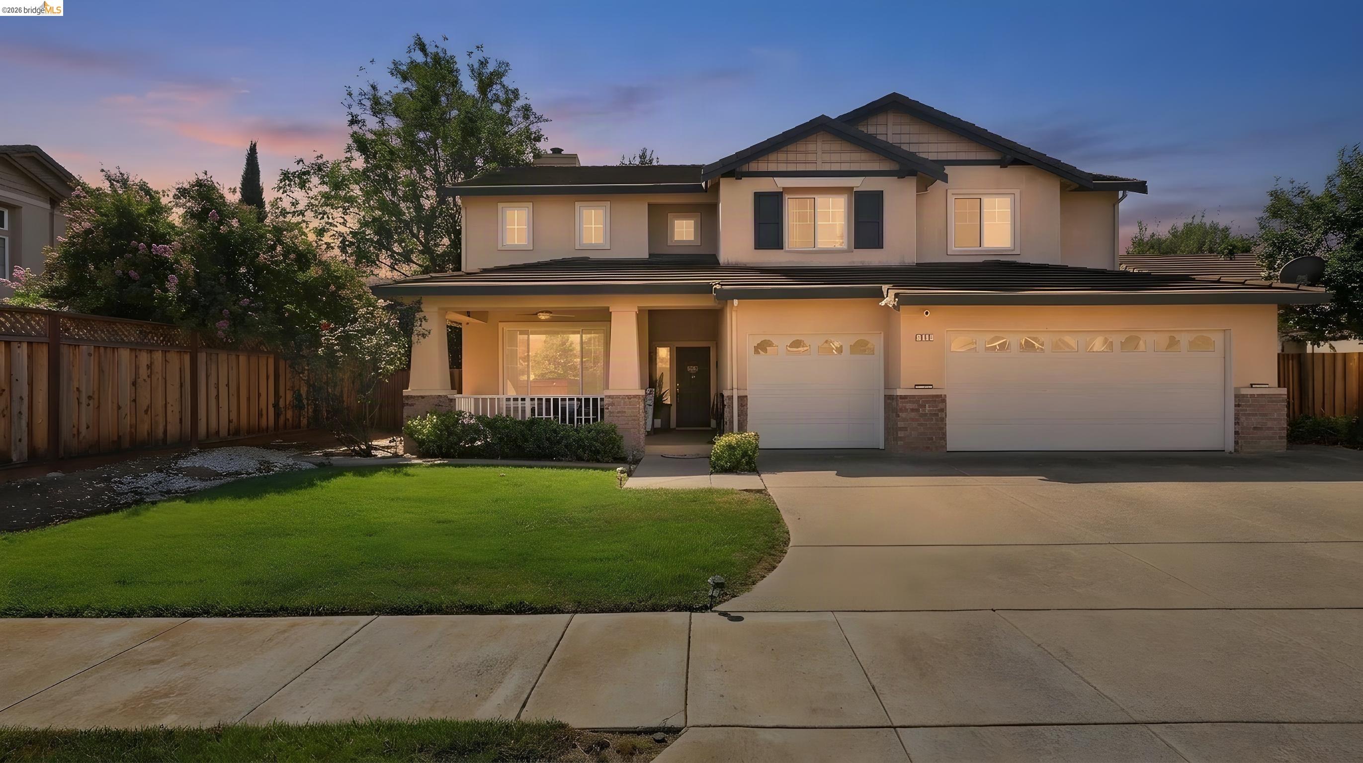 Two-story beige home with a covered porch, double garage, green lawn, and wide driveway at dusk.
