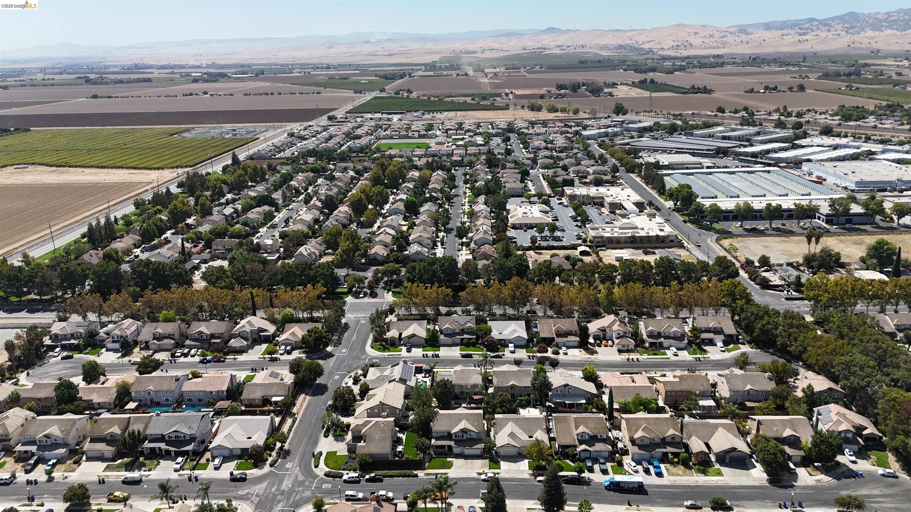 Aerial view of a suburban neighborhood bordered by farmland and industrial buildings, with dry hills in the distance.
