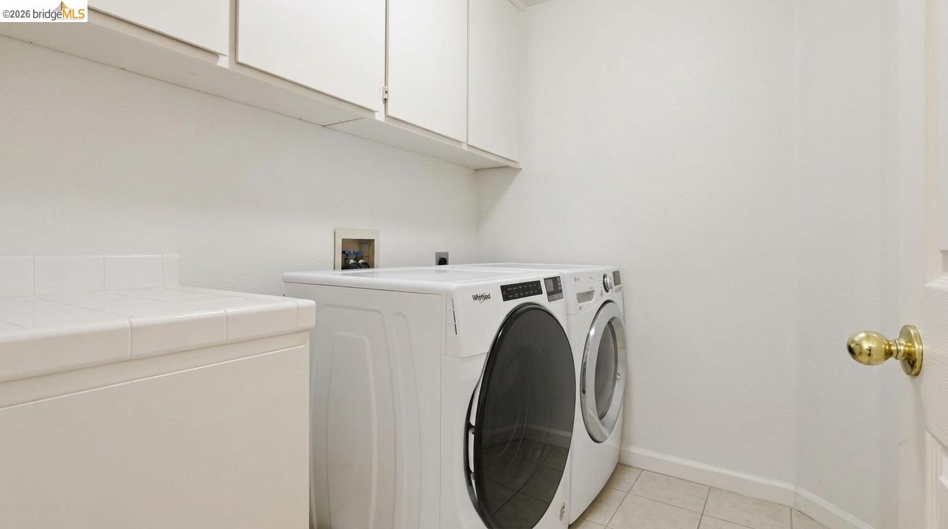 Laundry room with front-loading washer and dryer beneath white cabinets, beside a tiled utility sink.