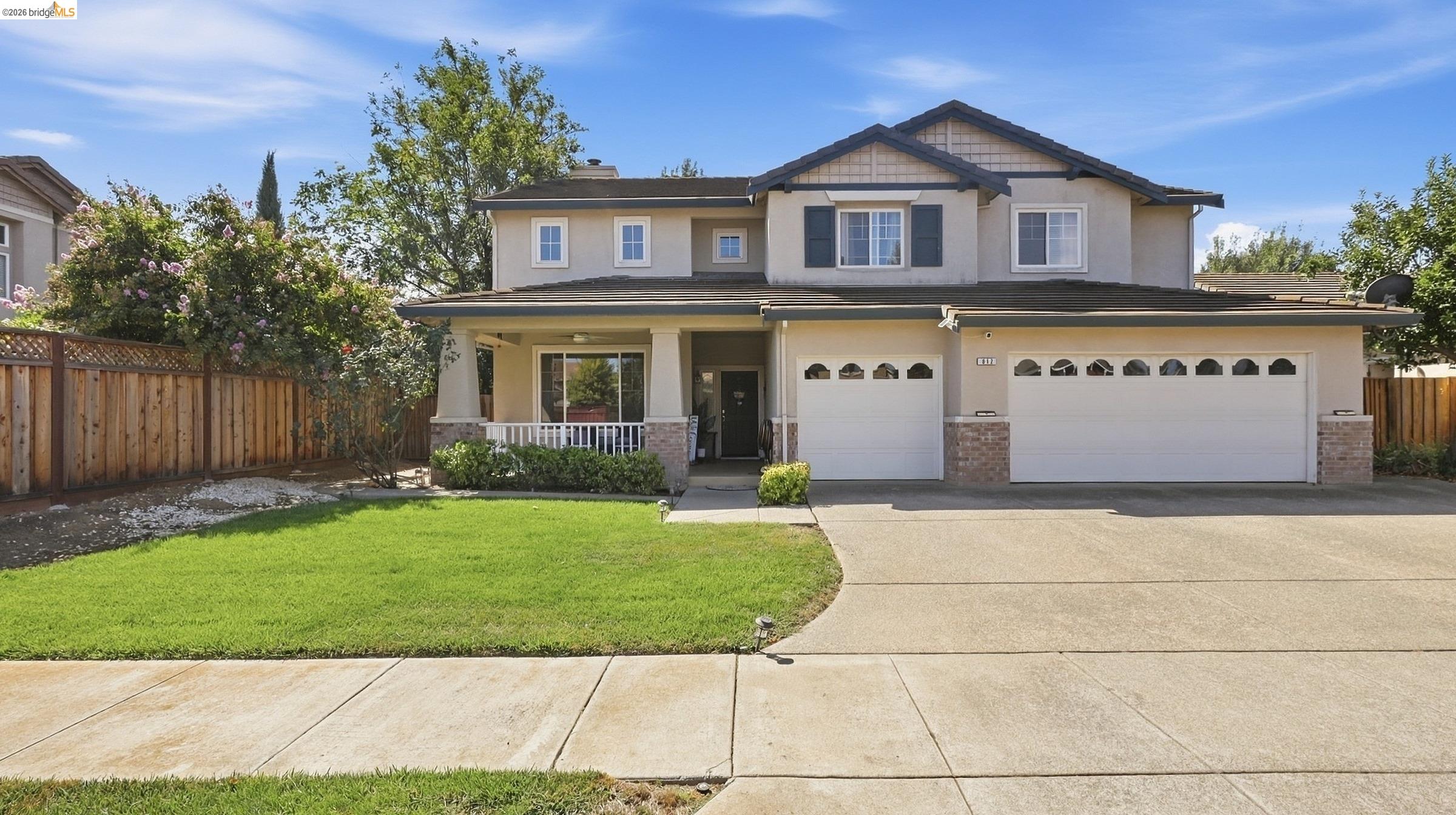 Two-story beige house with a covered porch, two-car garage, green lawn, and wide driveway.