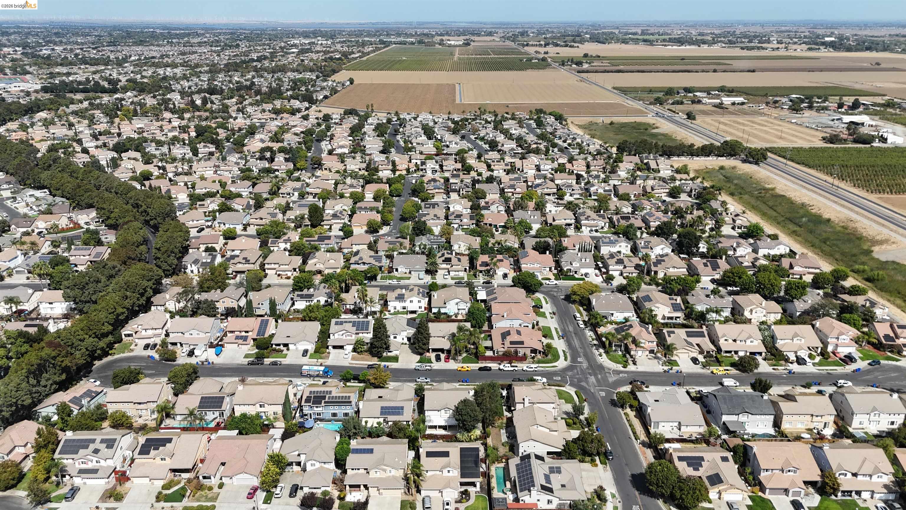 Aerial view of suburban homes and tree-lined streets beside expansive agricultural fields.