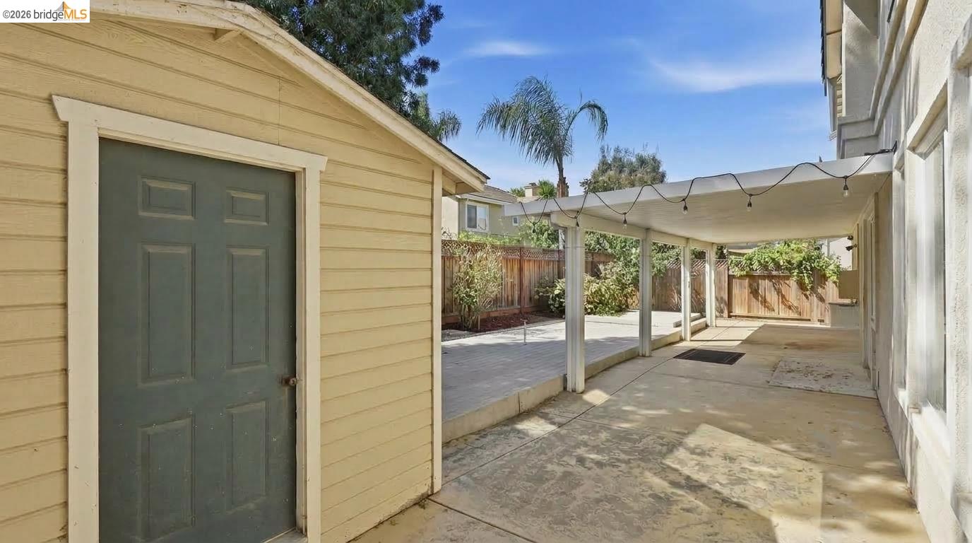 Backyard patio with a covered walkway, string lights, wooden fence, and small yellow shed.