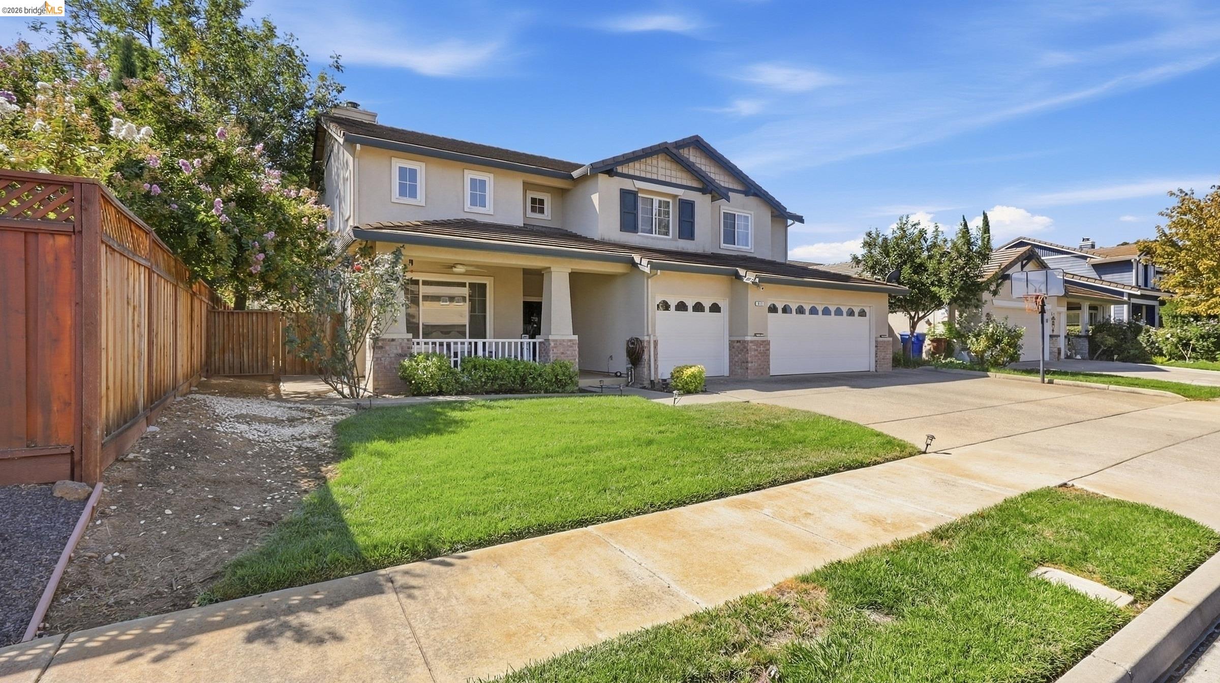 Two-story beige home with a covered porch, two-car garage, green lawn, and wooden side fence.
