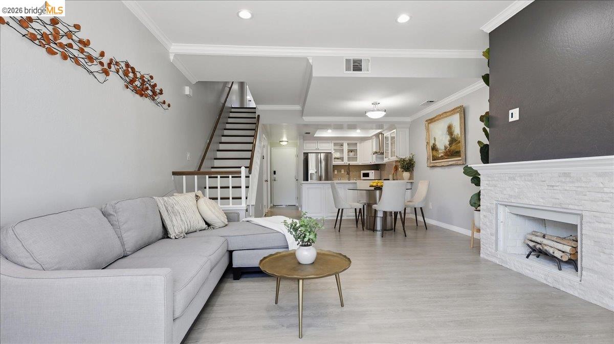 Staged living room with gray sectional, white brick fireplace, and open kitchen beyond.