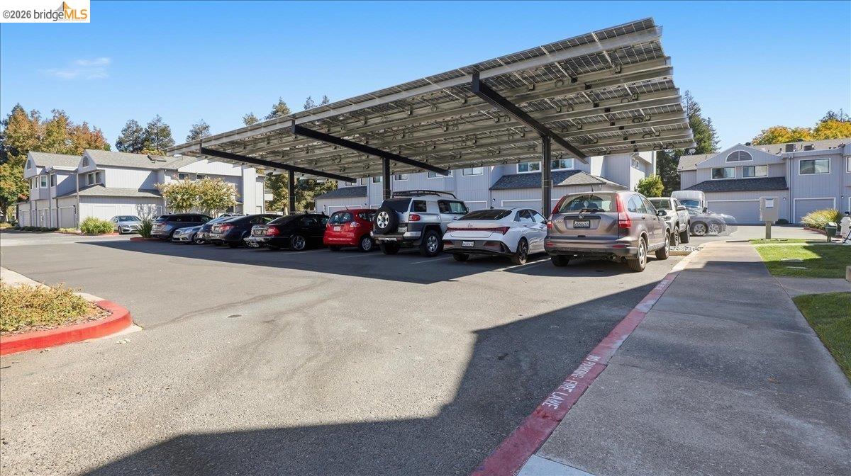 Residential parking area beneath a large solar-panel canopy, with parked cars and multi-unit buildings.