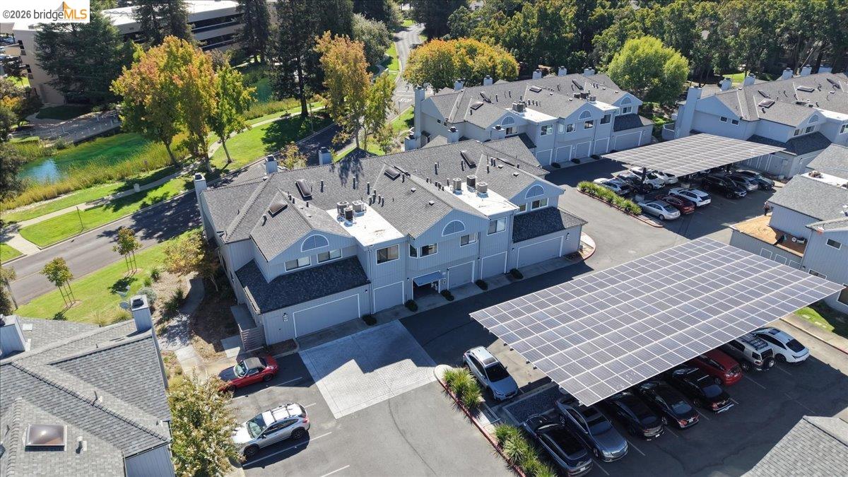 Aerial view of gray townhomes and solar-panel parking canopies beside a landscaped pond.