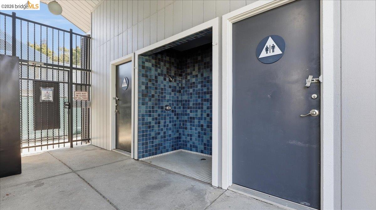 Outdoor pool-area shower with blue tile beside restroom doors and a gated walkway.
