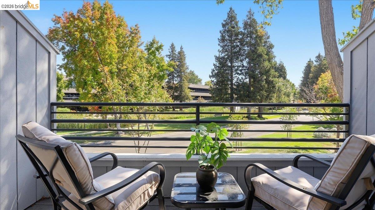 Furnished balcony with cushioned chairs and a potted plant overlooking a tree-lined street.