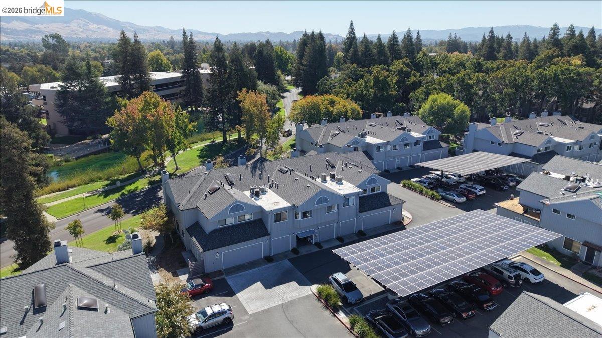 Aerial view of gray townhomes with a solar-panel parking canopy and wooded hills beyond.