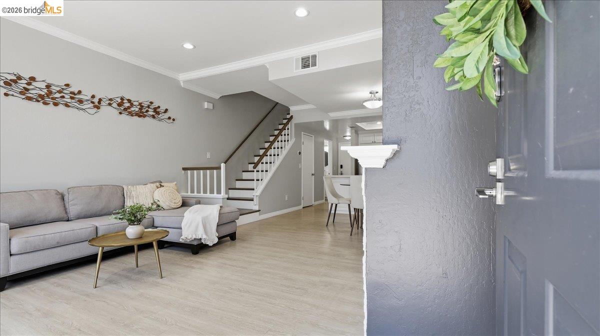 Entryway opens to a living room with a gray sectional, light wood floors, and a staircase.