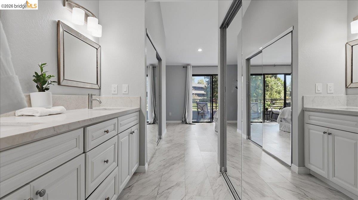 Bathroom with white paneled vanities, marble-look counters and flooring, and sliding mirrored closet doors.