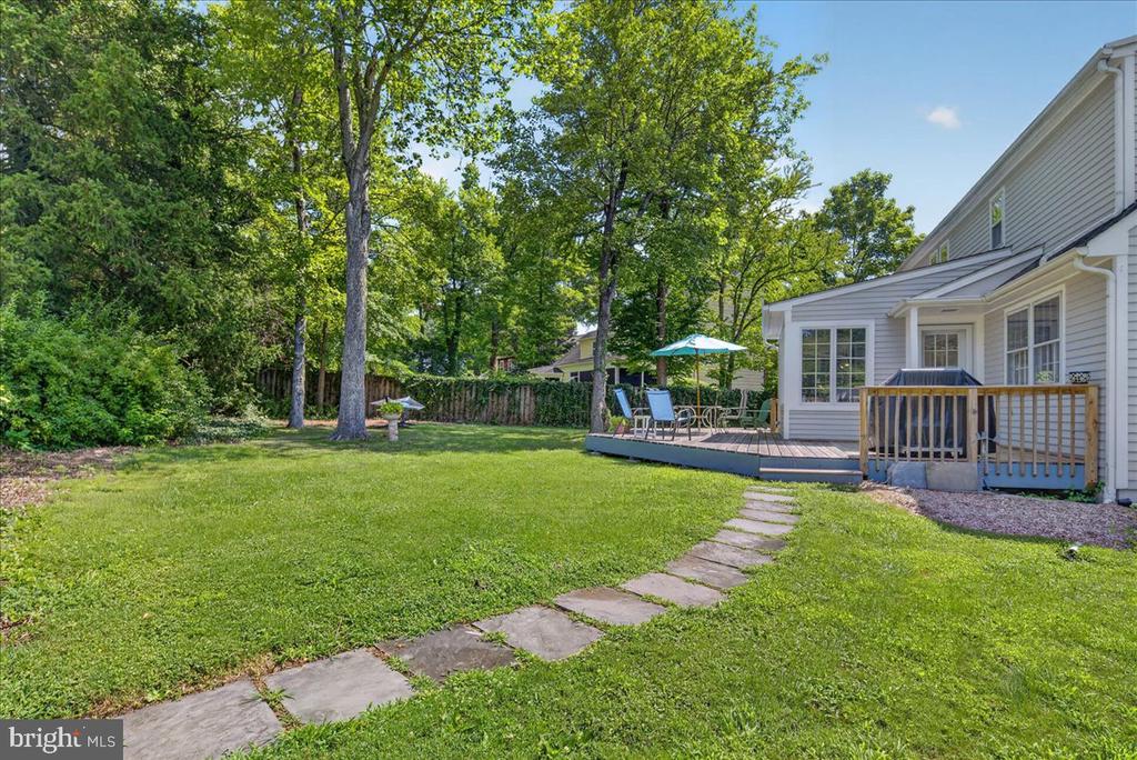 Backyard with broad lawn, stepping-stone path, wooded privacy, and furnished deck beside the house.