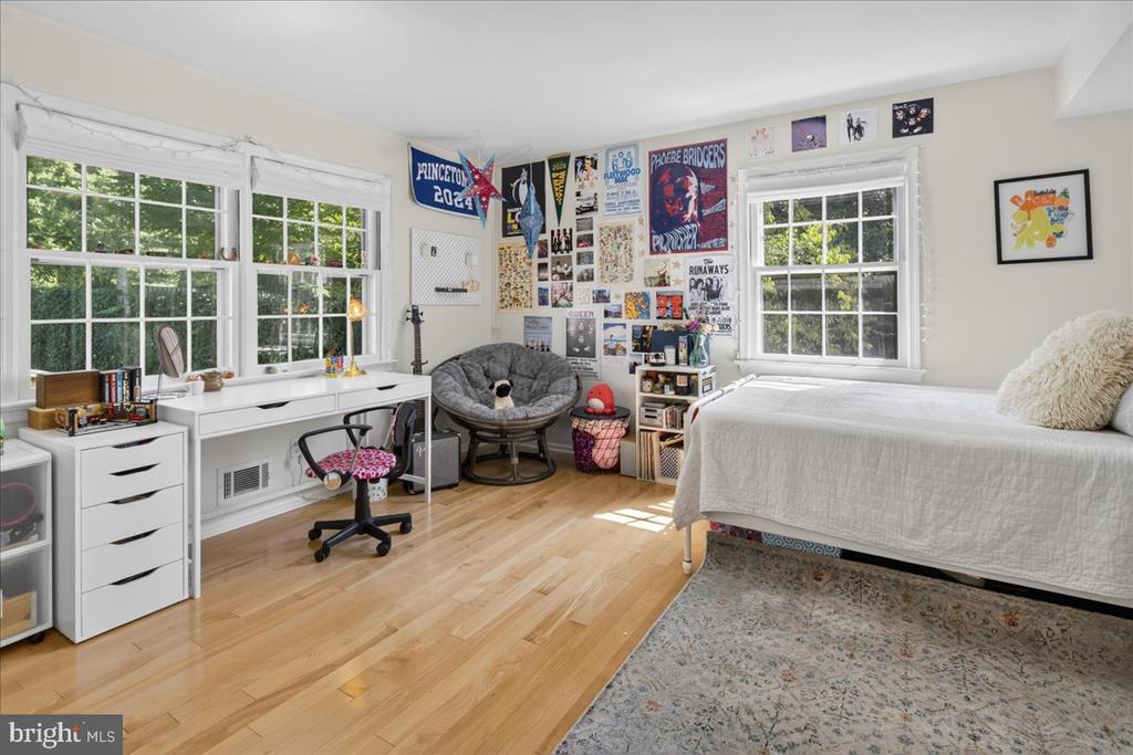 Bedroom with a white bed, large desk, hardwood floors, and a wall covered in colorful posters.