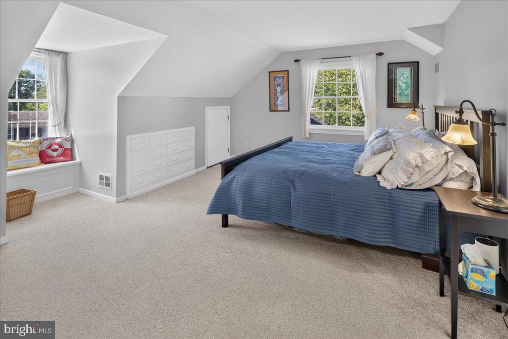 Attic bedroom with a blue-quilted bed, built-in dresser, and curtained dormer windows.