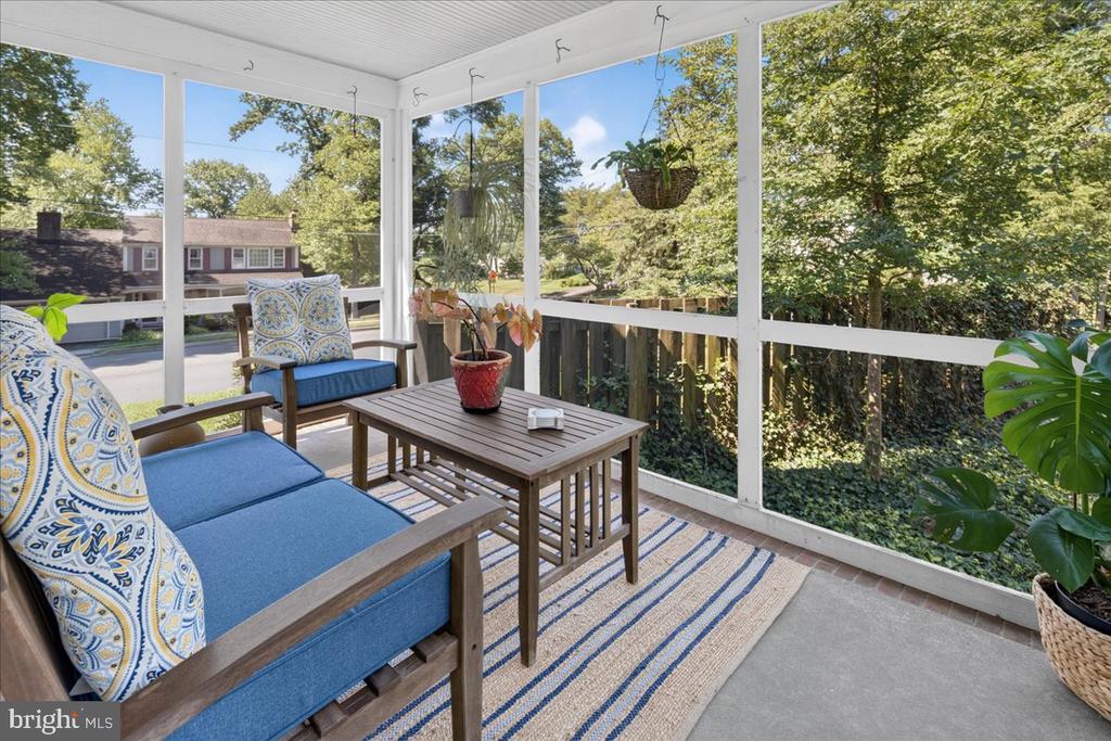 Glass-enclosed porch furnished with blue-cushioned chairs, a wooden table, and a striped rug.