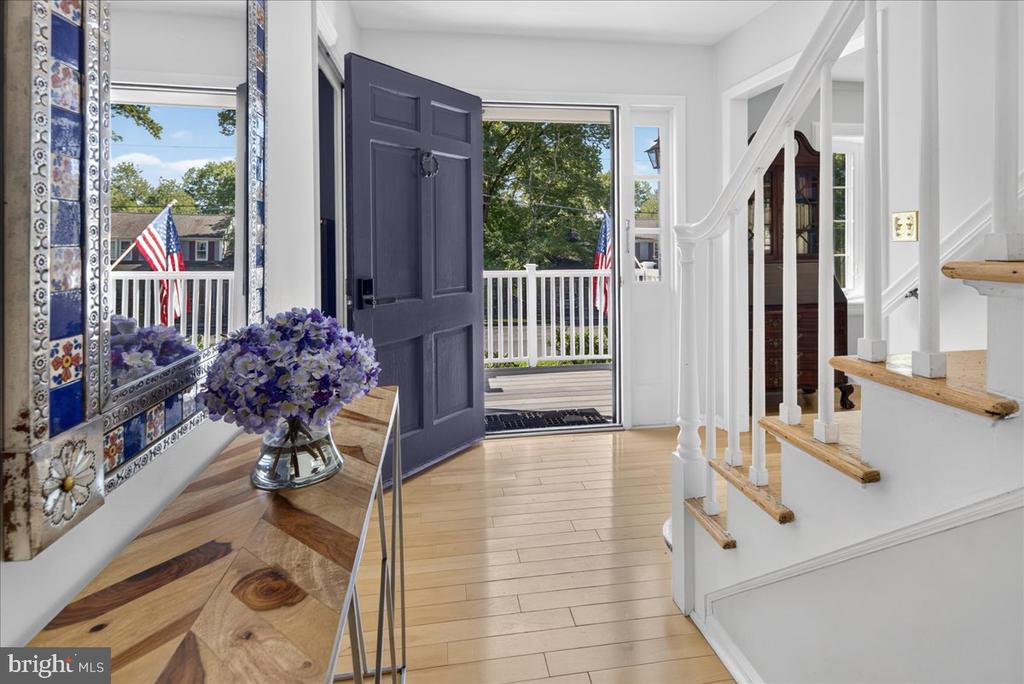 Entry foyer with a geometric wood console table, purple flowers, and an open front door beside a white staircase.