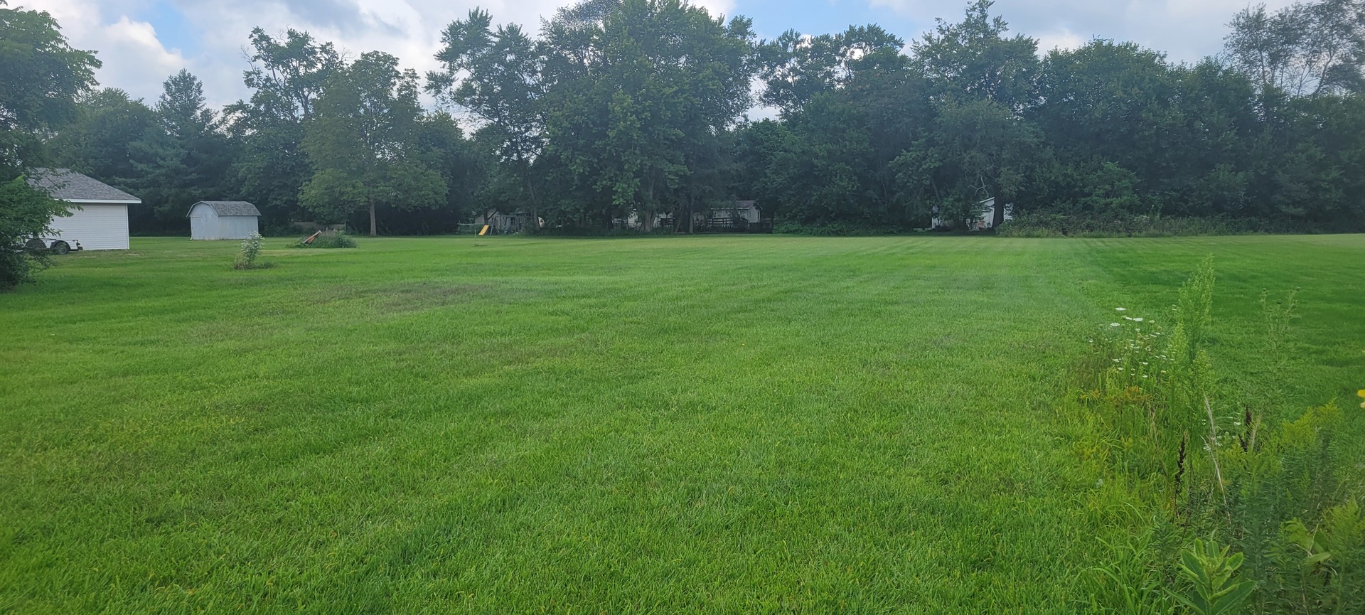 Wide grassy lot bordered by trees, with a small shed and play structures in the distance.