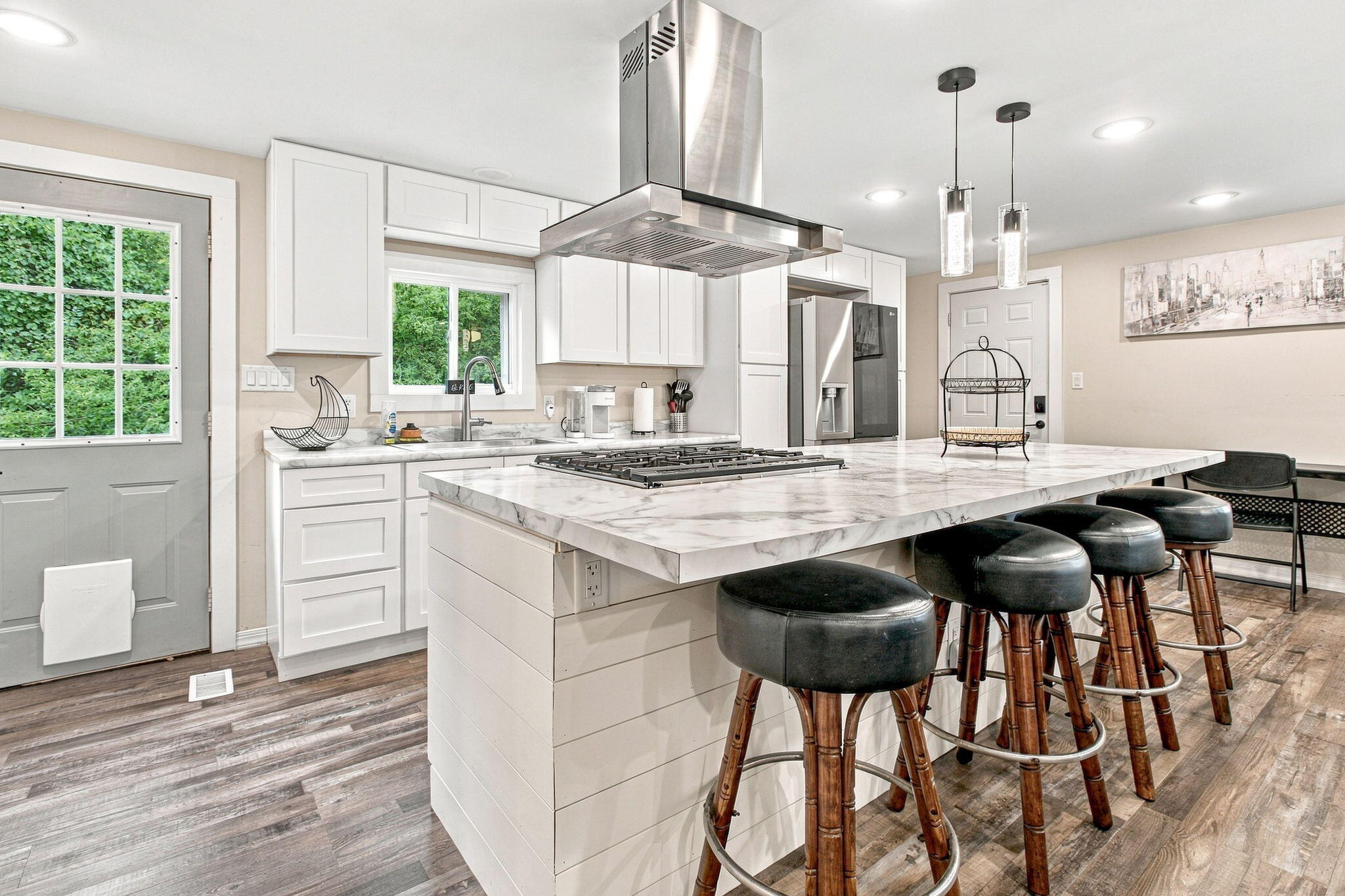 Modern kitchen with a large cooktop island, white cabinets, stainless steel appliances, and black bar stools.