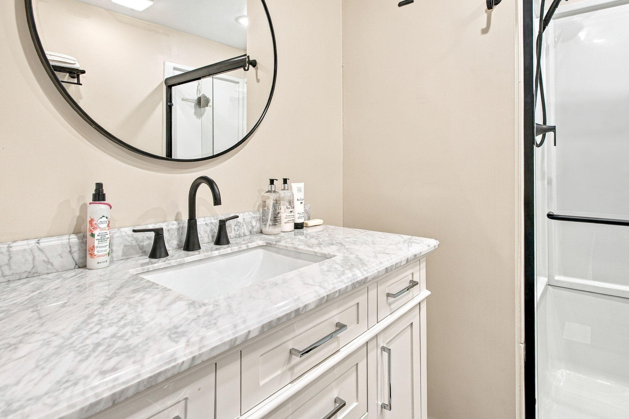 Bathroom vanity with marble counter, black fixtures, round mirror, and glass shower.