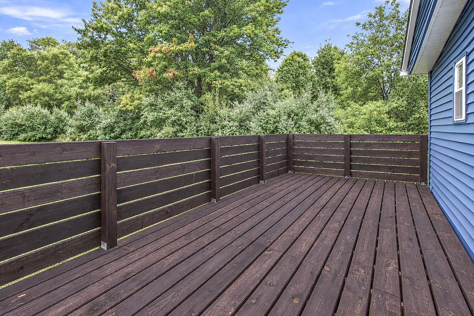 Wooden deck with dark privacy fencing beside a blue-sided house, surrounded by leafy trees.