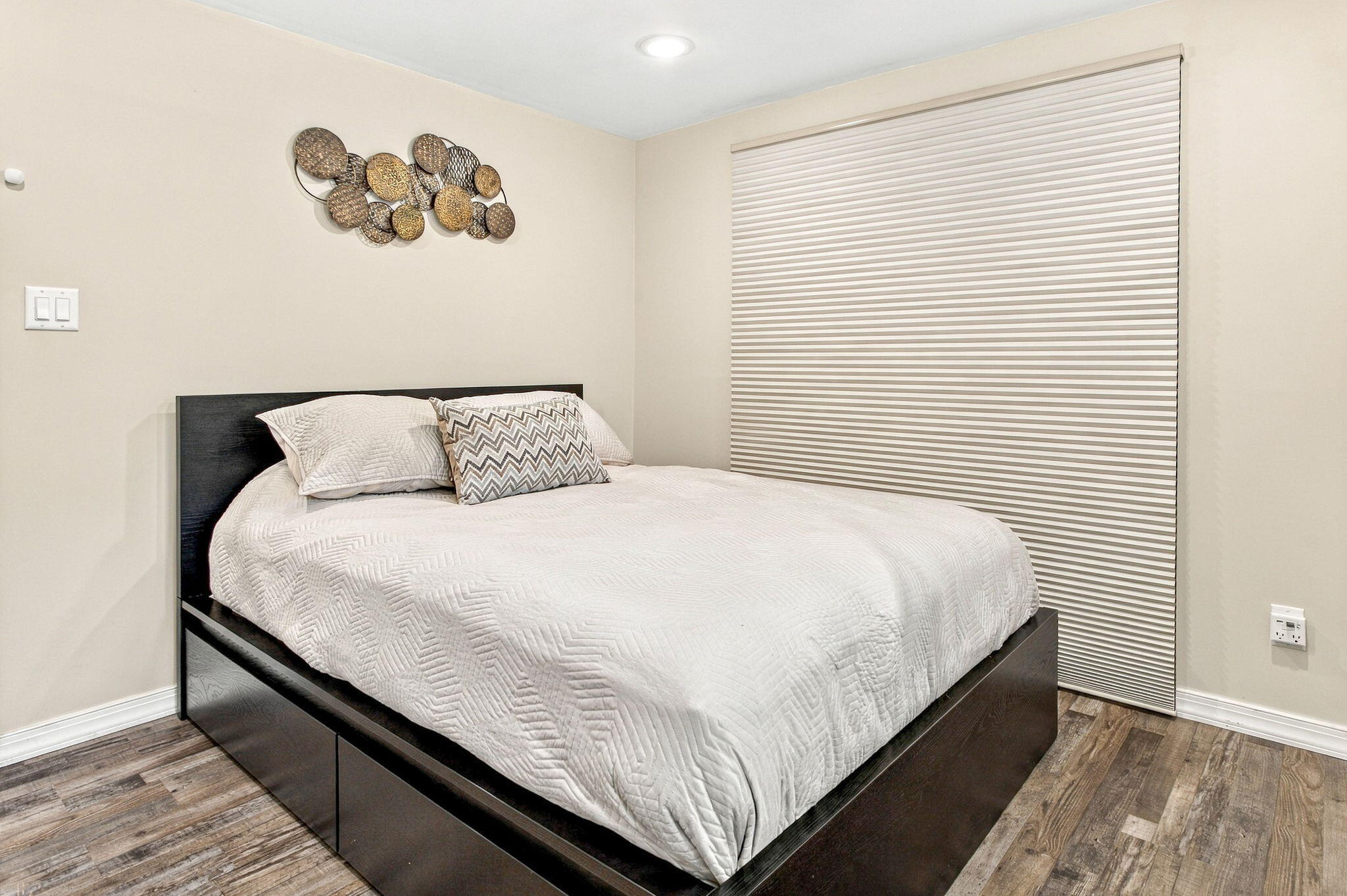 Bedroom with a dark platform bed, white quilted bedding, and a large striped window shade.