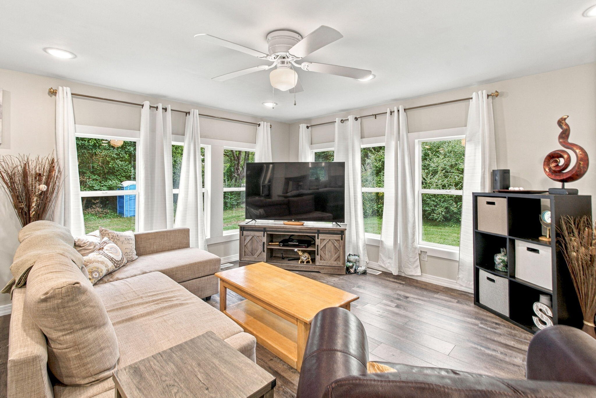 Living room with a beige sectional sofa, large TV, and windows overlooking greenery.