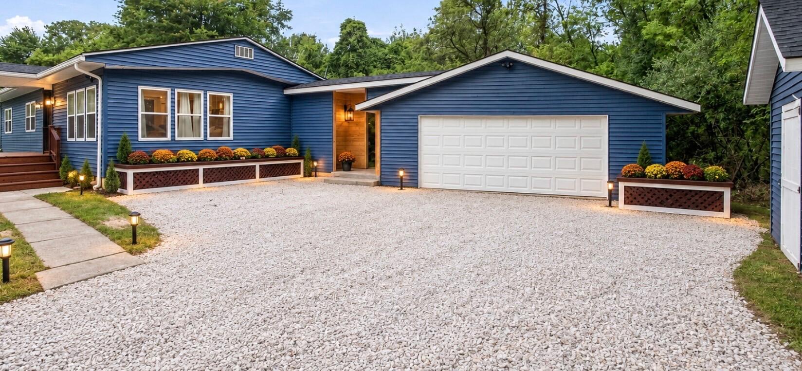 Blue home with attached white two-car garage, gravel driveway, raised flower beds, and walkway lights.