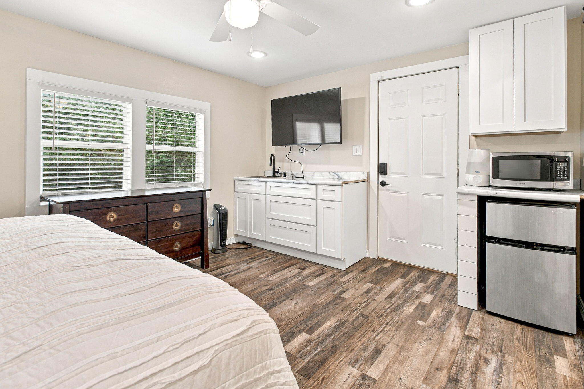 Bedroom with a compact kitchenette, wall-mounted TV, and two windows with white blinds.