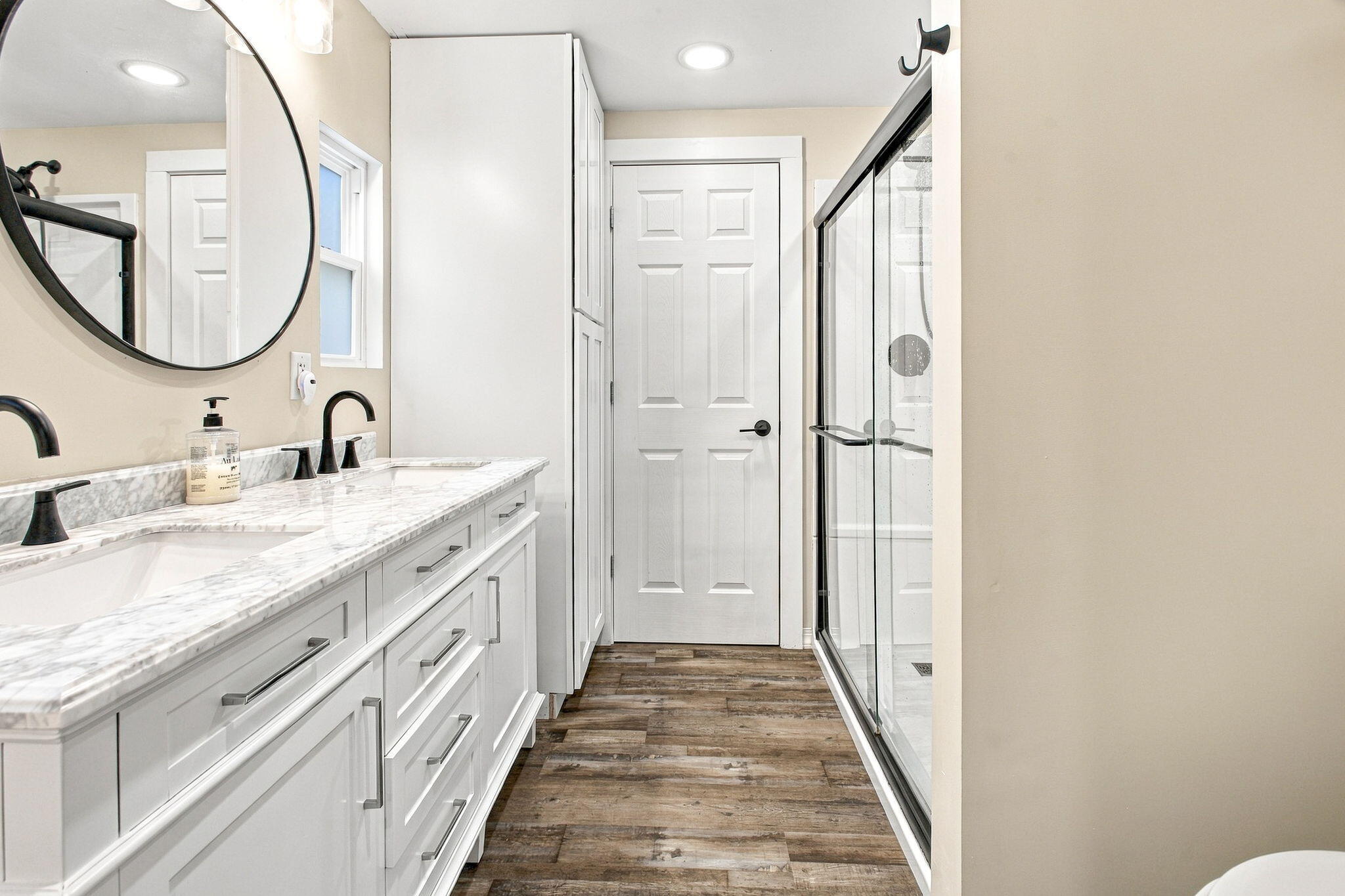 Bathroom with a long white vanity, marble-look counter, round mirror, and glass-enclosed shower.