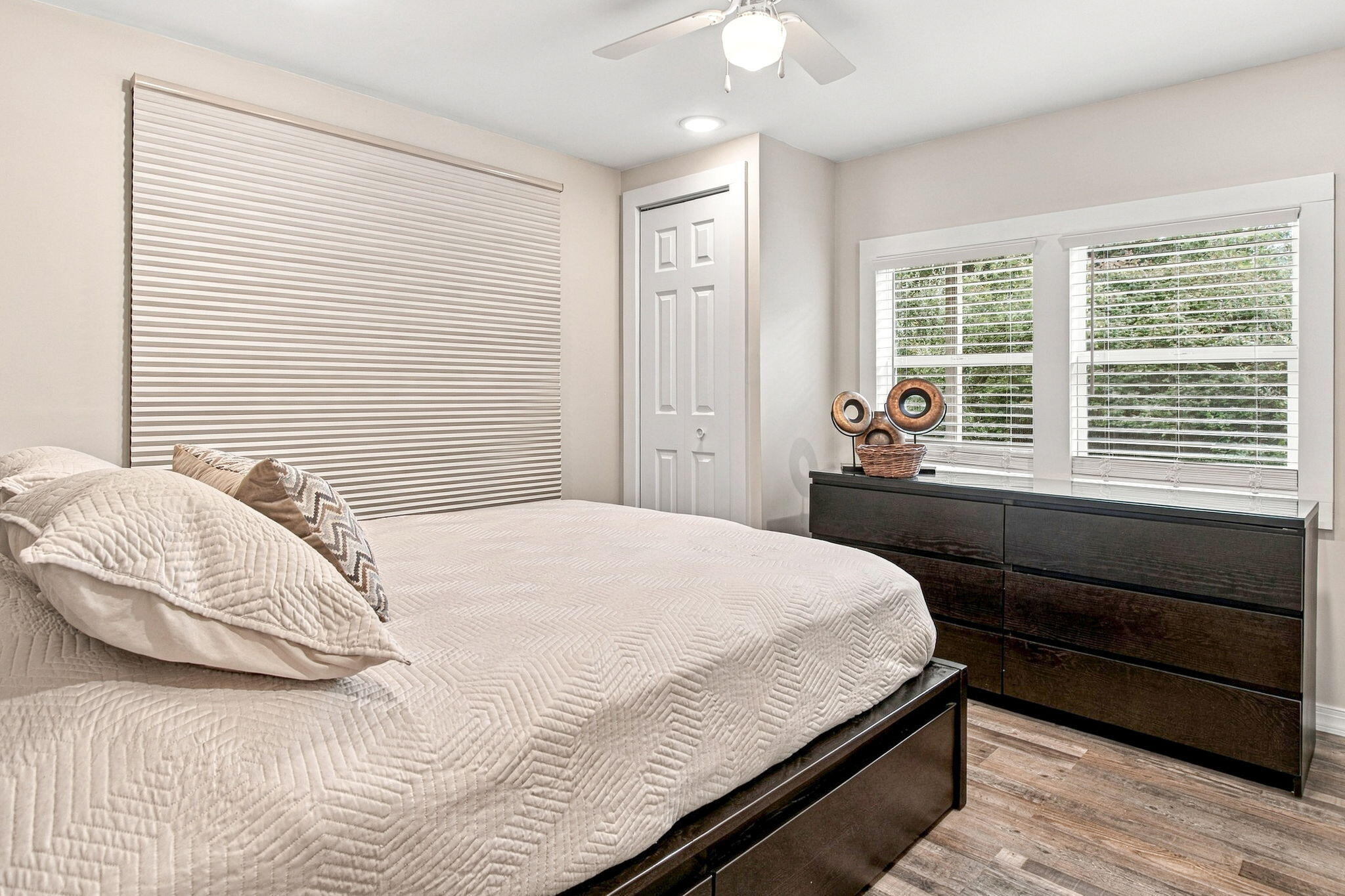 Bedroom with a white quilted bed, dark wood dresser, and two windows with blinds.