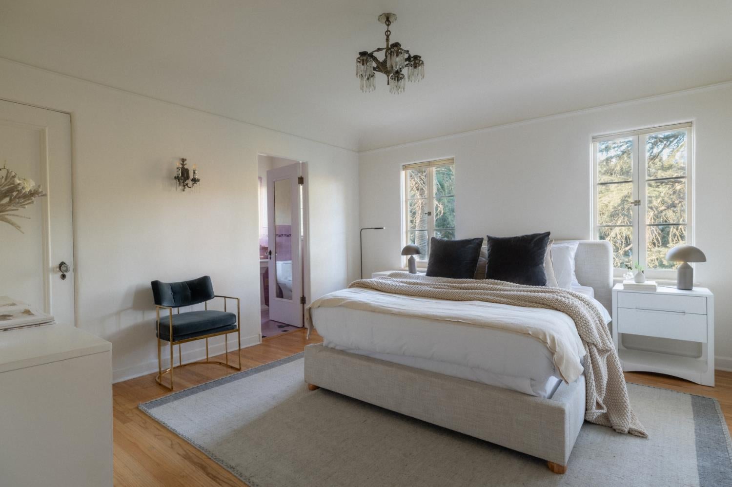 Bright bedroom with a large white bed, dark pillows, chandelier, and tall windows overlooking trees.
