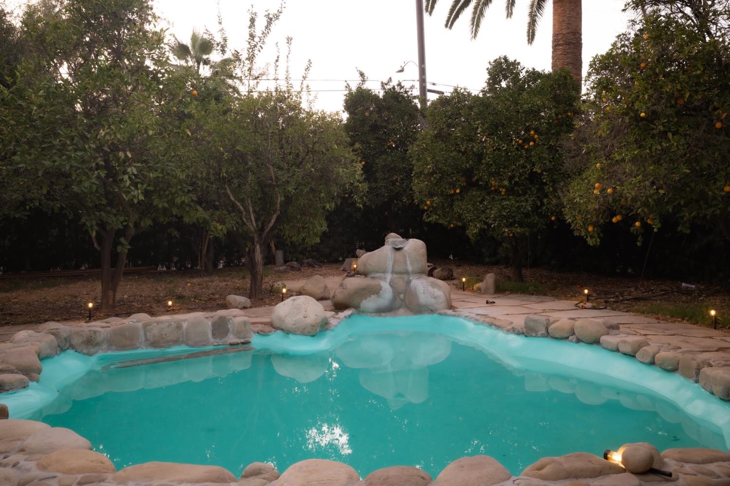 Backyard swimming pool with a stone waterfall, citrus trees, and glowing landscape lights.