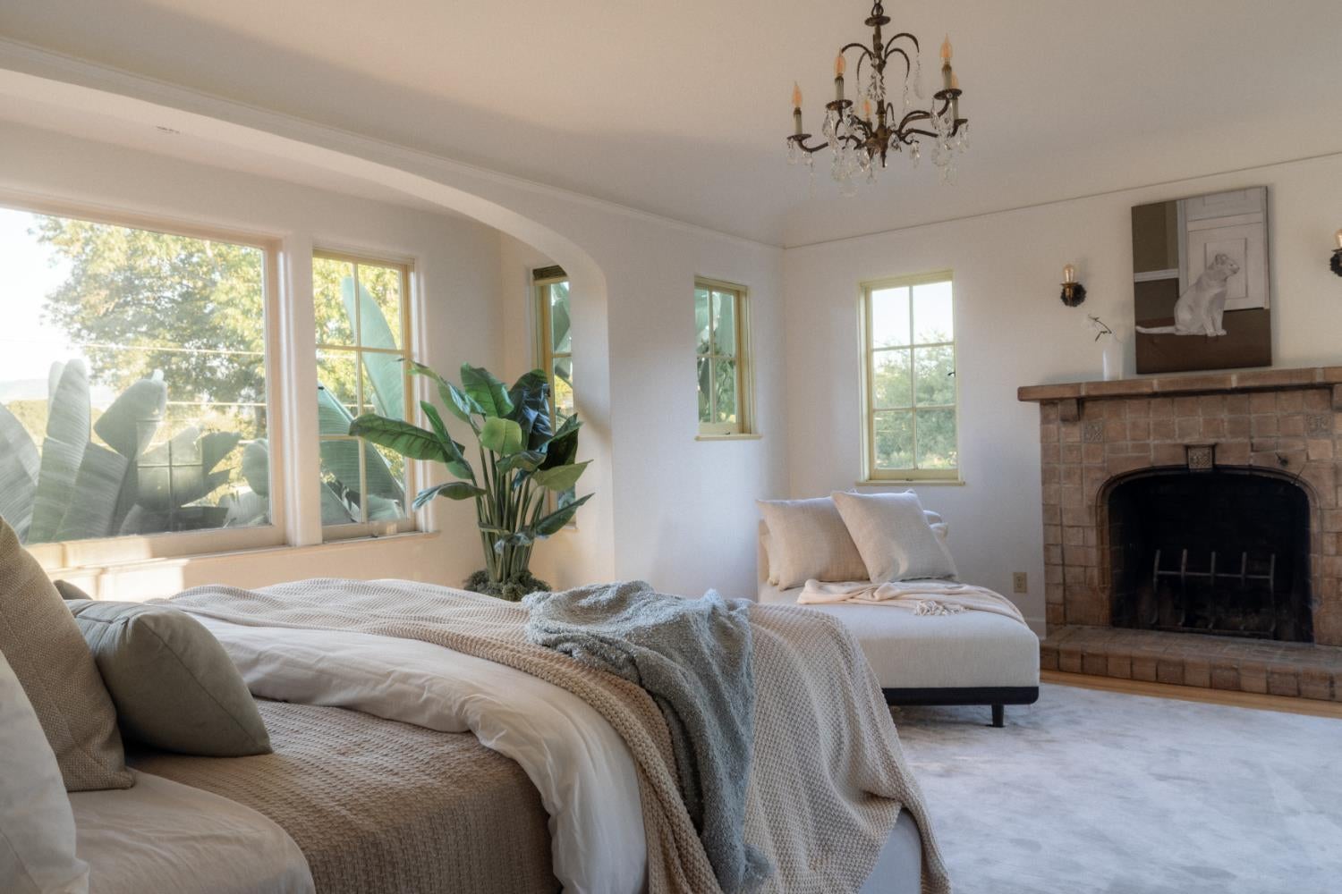 Bedroom with neutral layered bedding, tall windows, a tropical plant, and a brick fireplace.