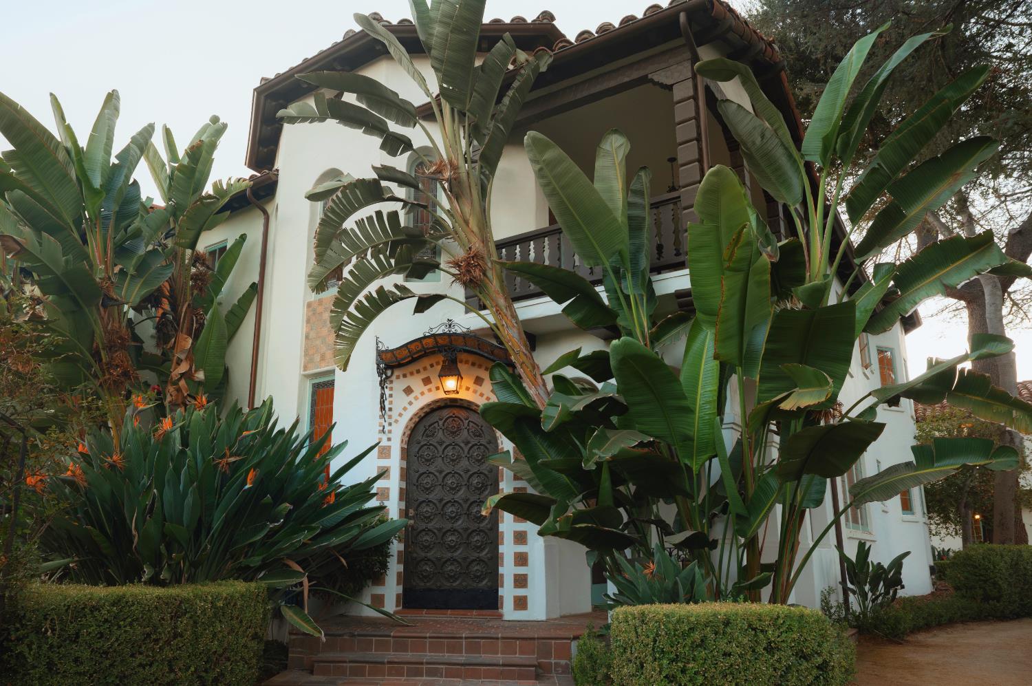 White stucco home with an ornate arched entry, second-floor balcony, and lush tropical landscaping.