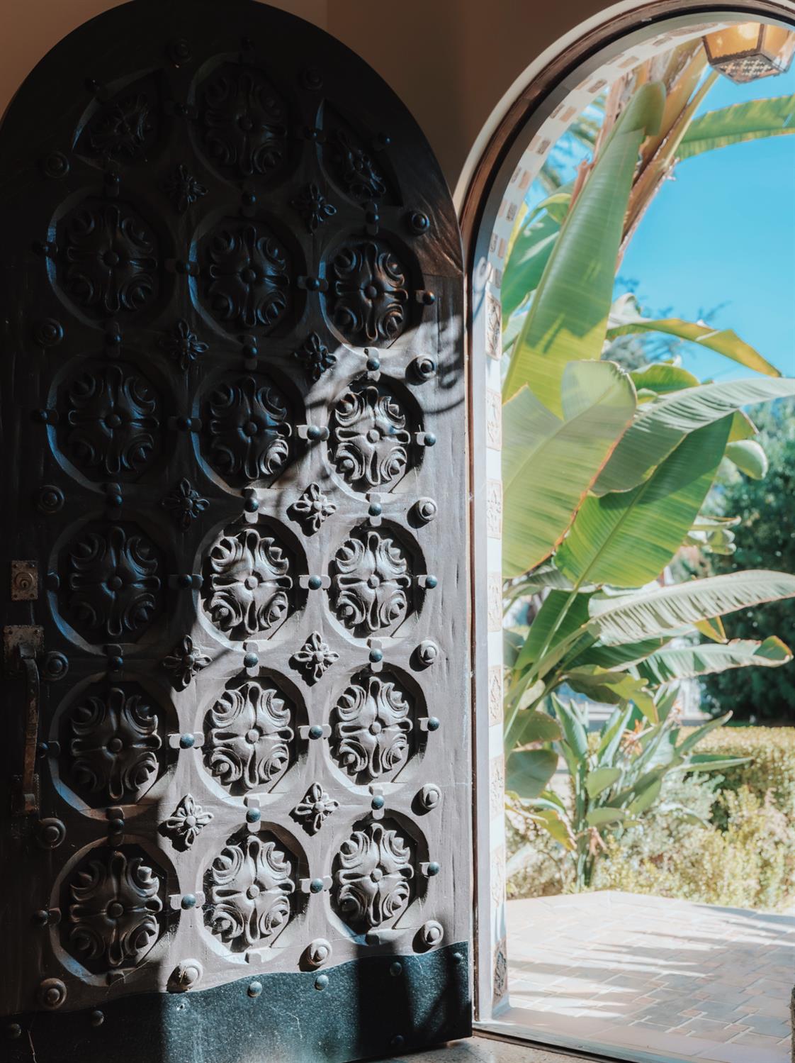 Ornately carved wooden entry door open to a sunlit patio with broad tropical leaves.