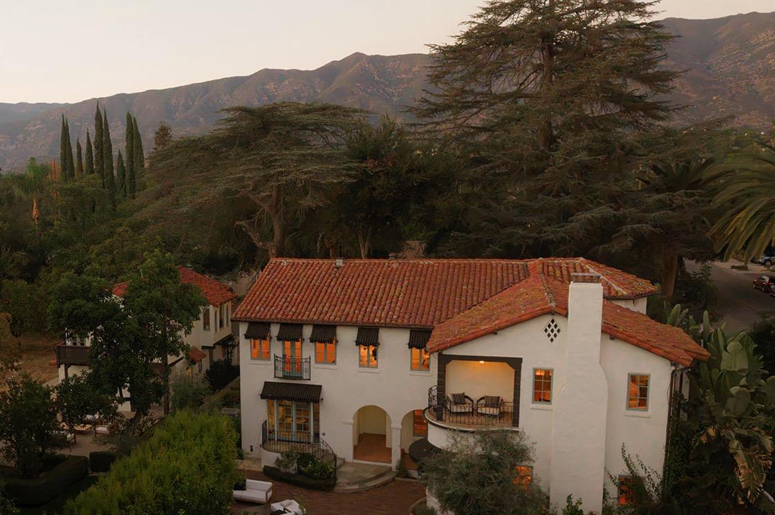 Two-story white stucco home with red tile roofs and balconies, framed by mature trees and mountain views.