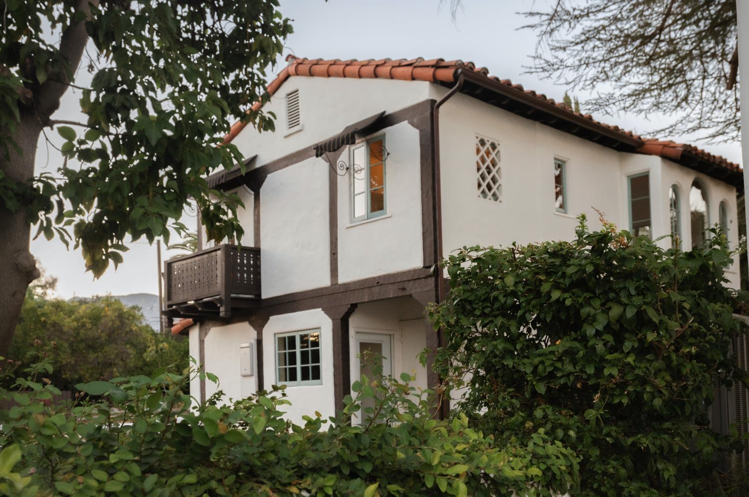 Two-story white stucco house with a red tile roof, dark trim, and a small upper balcony behind leafy hedges.